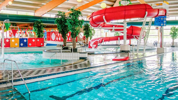 An indoor swimming pool with a red water slide, multiple lanes, and a lifeguard stand.
