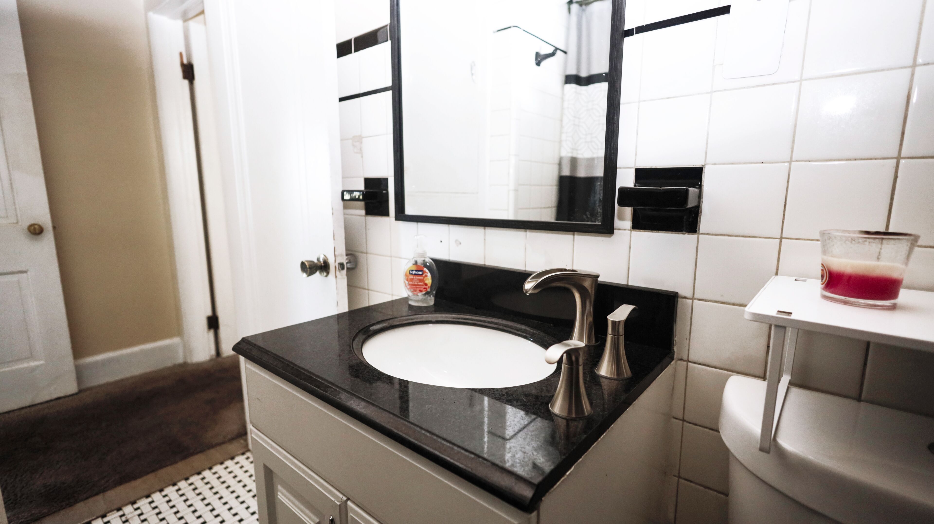 Une salle de bain avec un lavabo blanc, un plan de toilette noir et un miroir au-dessus. On y trouve un flacon de savon liquide sur le plan de toilette et un verre de boisson rouge sur une petite étagère blanche.