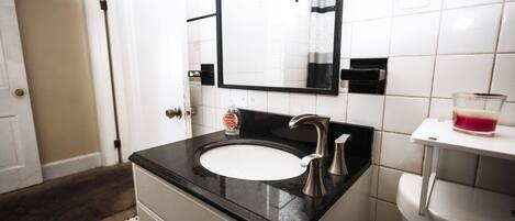 Une salle de bain avec un lavabo blanc, un plan de toilette noir et un miroir au-dessus. On y trouve un flacon de savon liquide sur le plan de toilette et un verre de boisson rouge sur une petite étagère blanche.