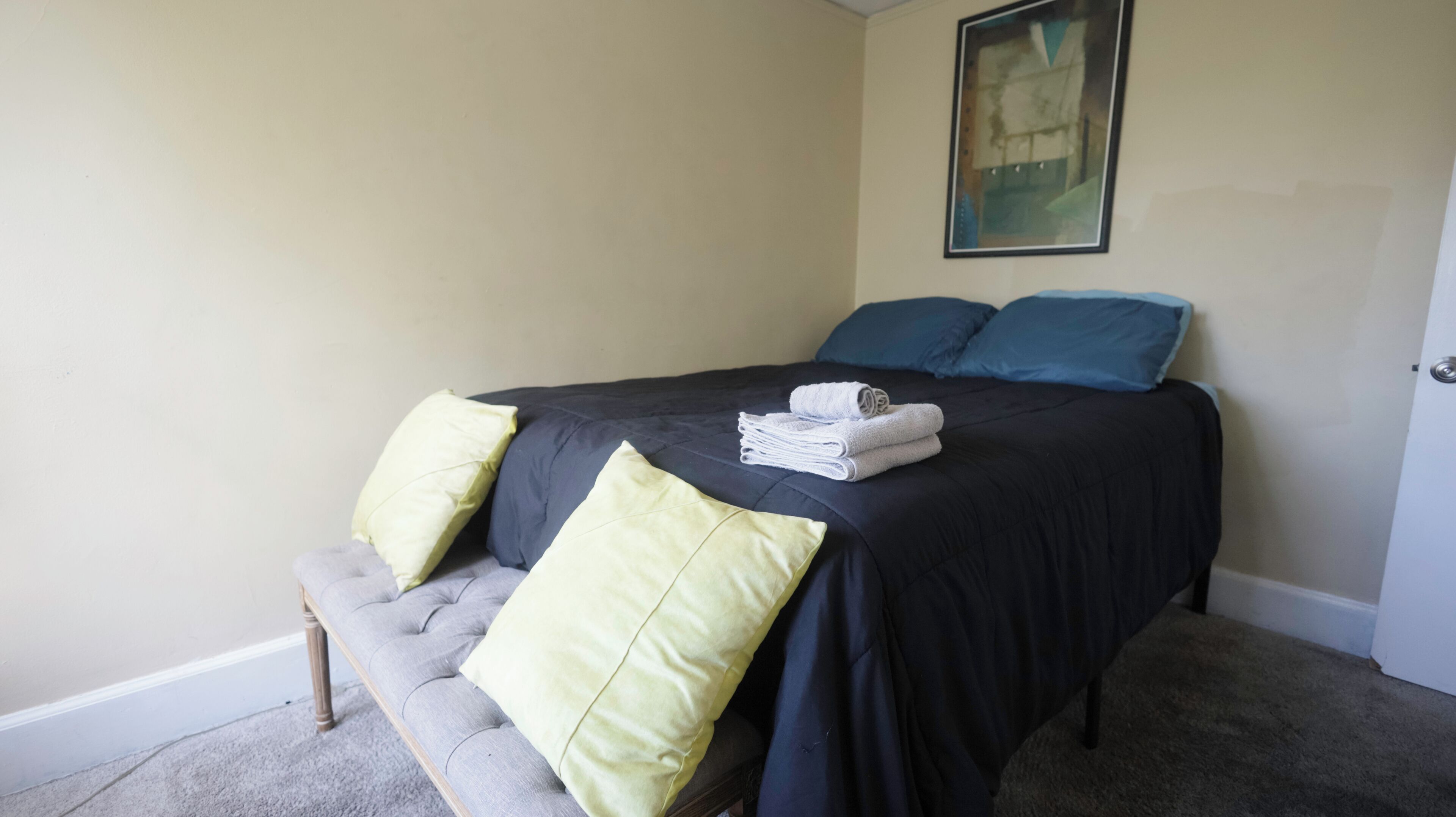 A neatly made bed with a dark bedspread, two pillows, and a stack of folded towels.
