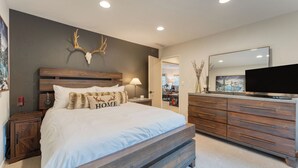 A bedroom with a large bed, wooden headboard, a mounted deer skull, a dresser, a television, and a painting on the wall. - Westlake Cabin by Avantstay Hot Tub, Near Skiing, Walk to Restaurants & Lake! (Tahoe City)
