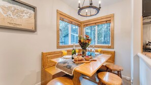 A dining area with a wooden table, bench seating, a chandelier, and a window with a view of trees. - Westlake Cabin by Avantstay Hot Tub, Near Skiing, Walk to Restaurants & Lake! (Tahoe City)