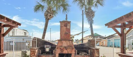 An outdoor barbecue area with a stone-built grill, surrounded by wooden benches and palm trees.