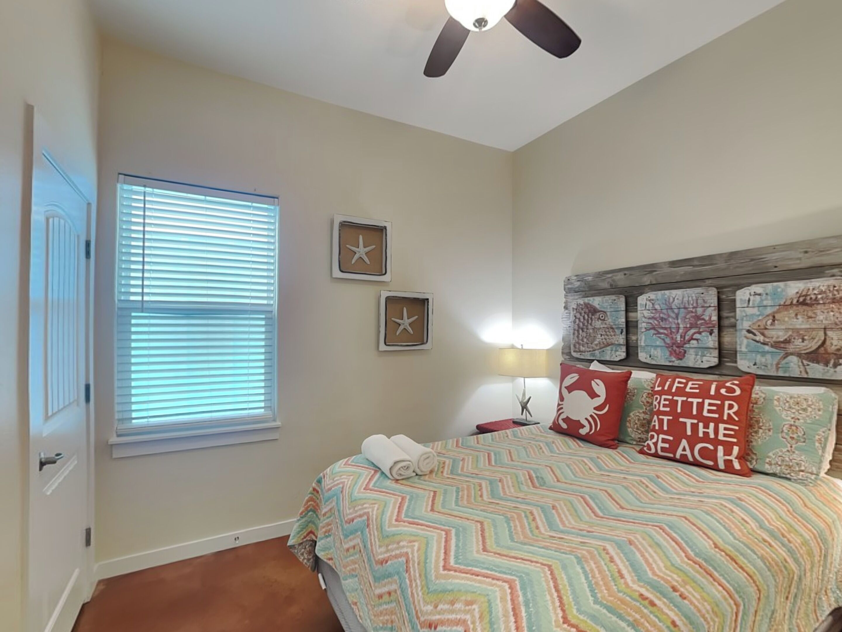 Une chambre à coucher comprenant un lit, un ventilateur de plafond, une fenêtre avec des stores et des éléments de décoration murale, notamment des étoiles de mer et des poissons.