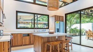 A modern kitchen with wooden cabinetry, a large island with bar stools, a built-in oven, and a large window overlooking a garden.