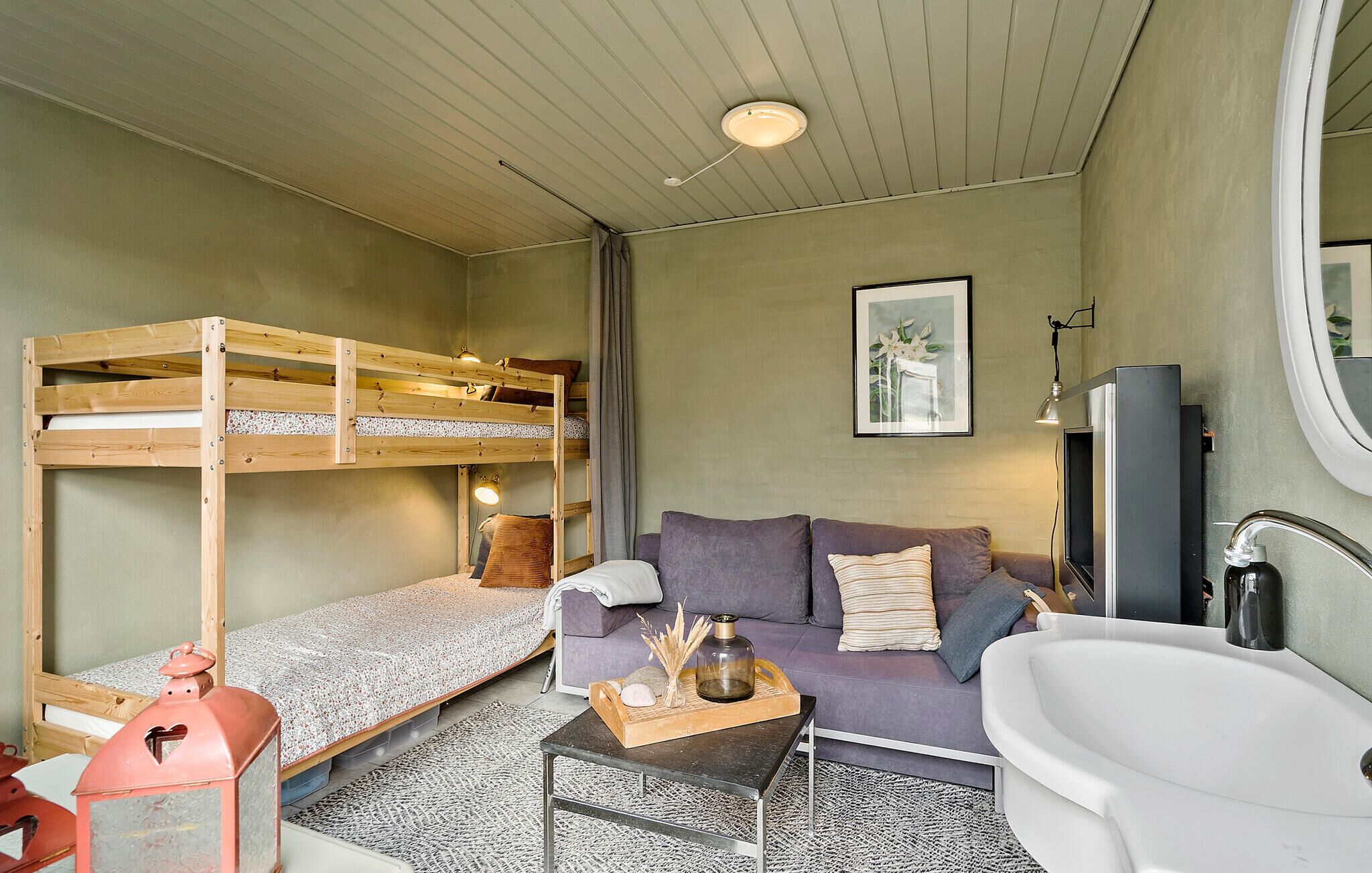 Une petite pièce comprenant un lit superposé, un canapé, une petite table agrémentée d’objets décoratifs, un lavabo et un tableau encadré au mur.