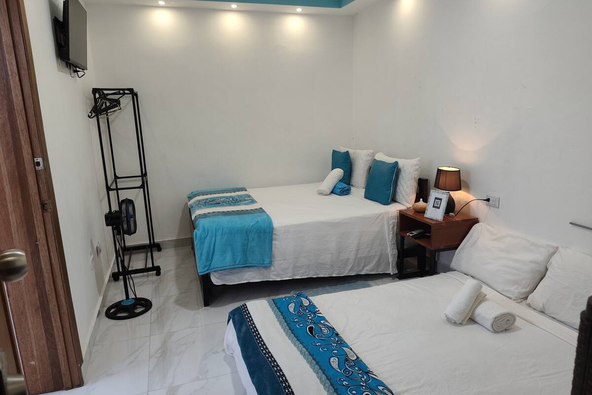 A hotel room with two beds, a TV mounted on the wall, a bedside table with a lamp, and a fan on the floor.