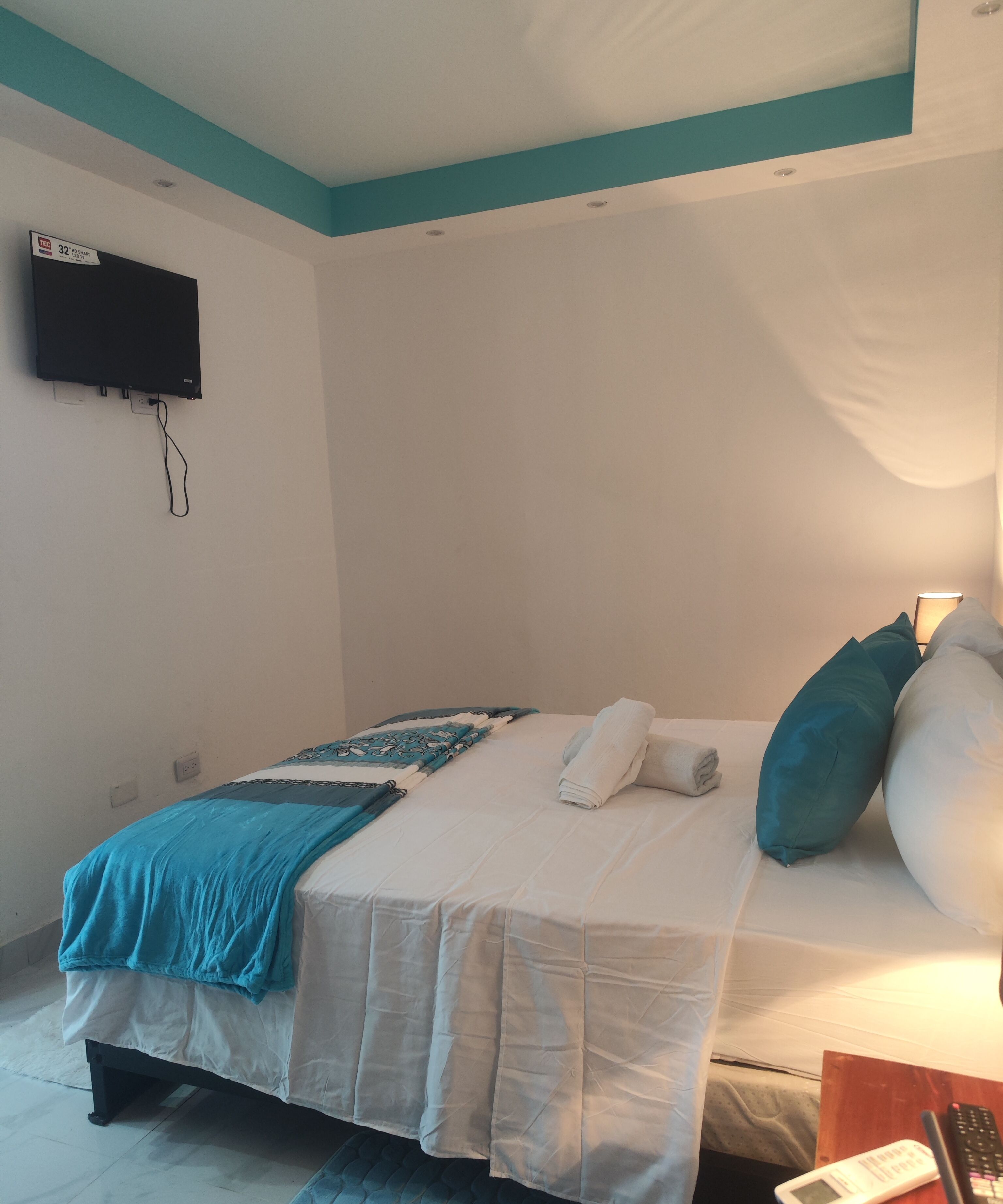 A bedroom with a bed, a TV mounted on the wall, a bedside table with a lamp, and a remote control on the table.
