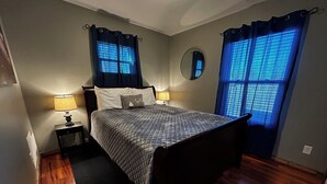 Un dormitorio con cama, dos mesitas de noche con lámparas, un espejo redondo y una ventana con cortinas azules.