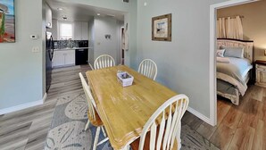 A dining area with a wooden table and chairs, a kitchen in the background, and a bedroom with a bed and curtains. - Coastal Hideaway by Silver Sands Vacation Rentals (Port Aransas)
