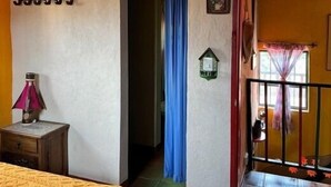 A room with a bed, a nightstand, a lamp, a door with a blue curtain, a small wall-mounted light, and a window with a curtain.