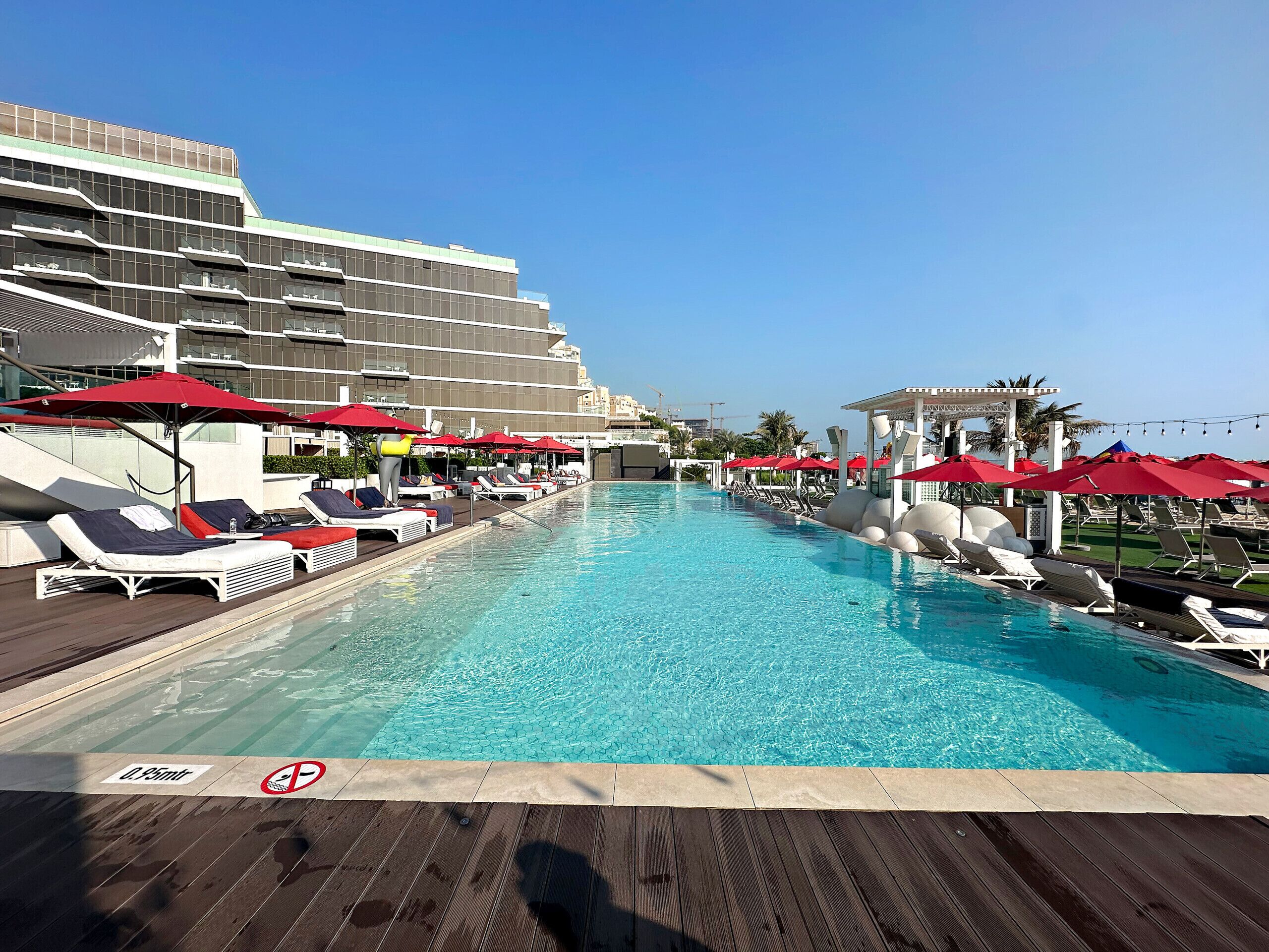 A swimming pool with sun loungers and red umbrellas, surrounded by a modern building.