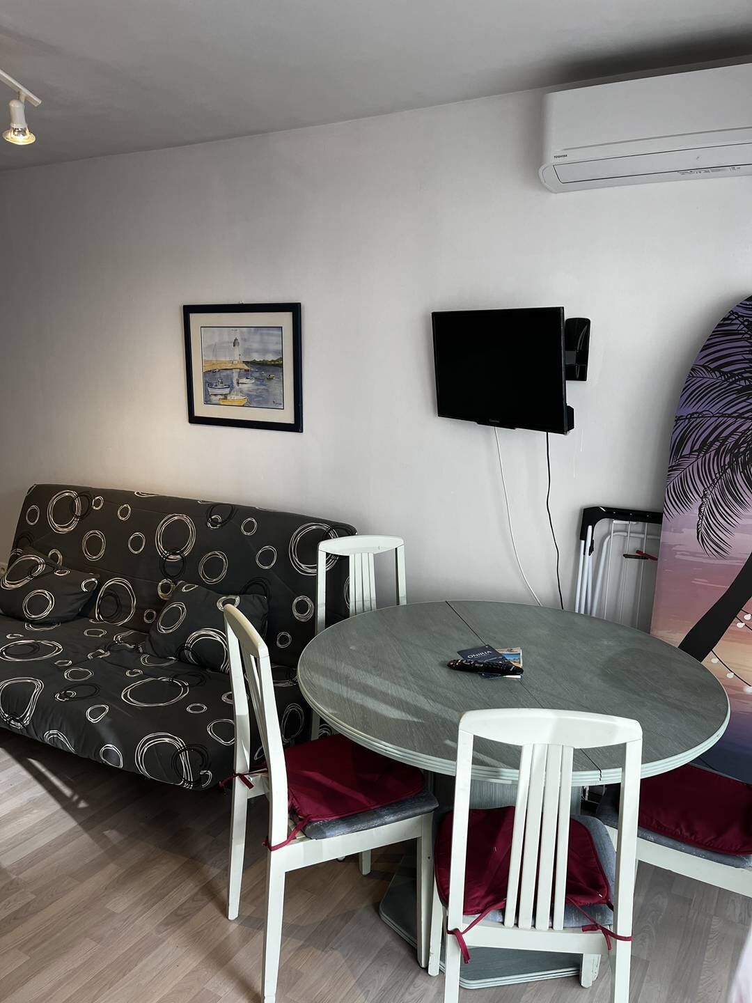 A modern living room with a round dining table, white chairs, a black sofa with a circular pattern, a framed picture on the wall, and a mounted TV.
