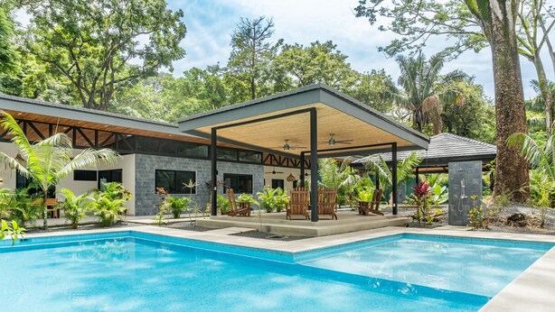 Un espace piscine moderne doté d’un coin salon abrité, entouré d’une végétation luxuriante et d’arbres.