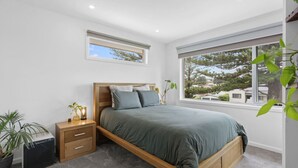 A modern bedroom with a wooden bed, a small bedside table, a potted plant, and a large window offering a view of trees and houses. - 3 bedroom, pet friendly home CBD (Warrnambool)