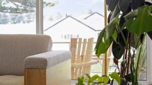 A room with a grey sofa, a wooden chair, and a large window offering a view of a house and greenery outside. - 3 bedroom, pet friendly home CBD (Warrnambool)