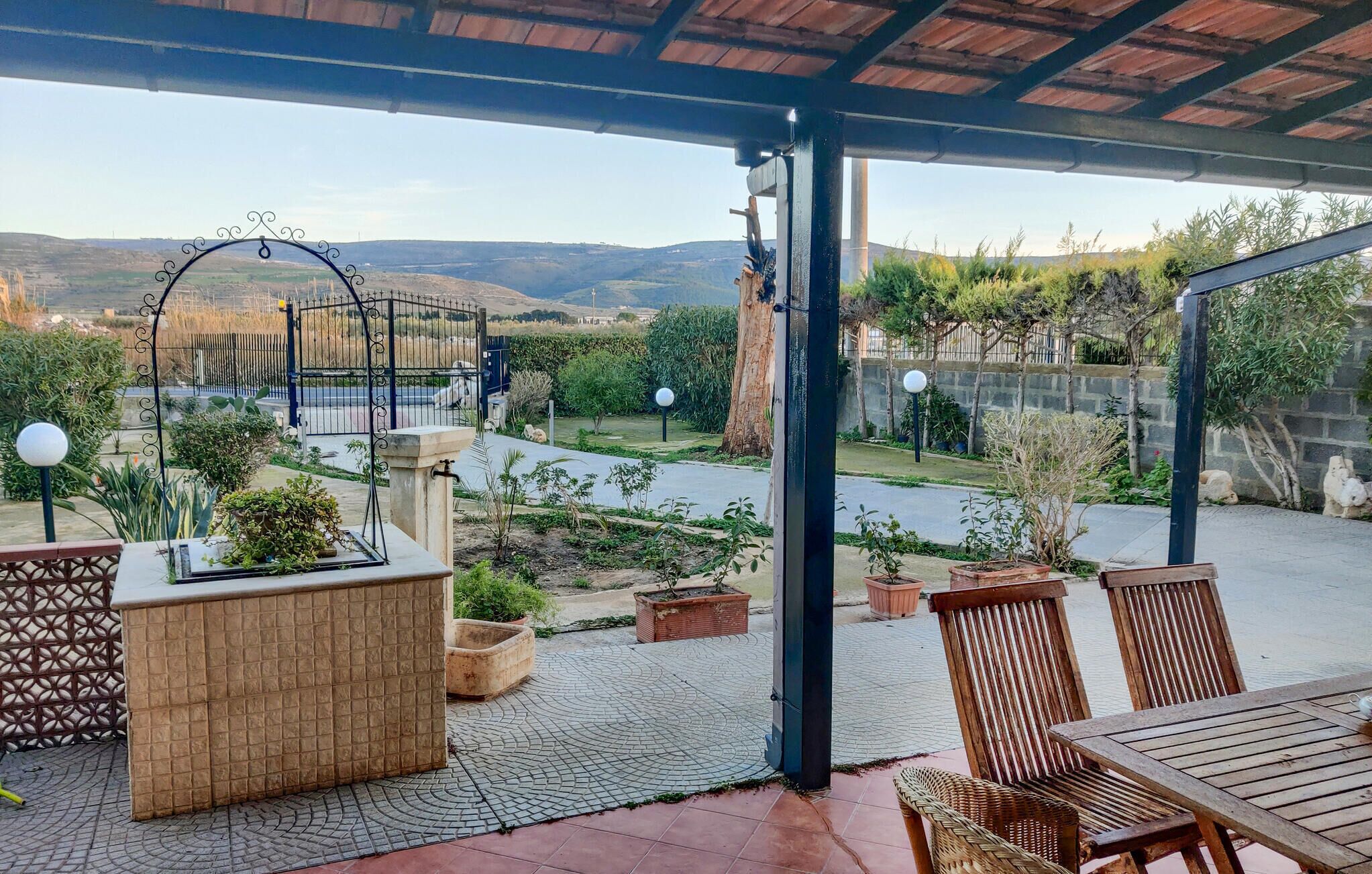 Un'area patio esterna con una zona salotto coperta, un vaso in pietra e vista su un giardino curato e dolci colline in lontananza.