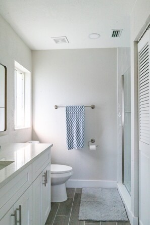 A bathroom with a toilet, a towel rack with a patterned towel, a shower area, and a window providing natural light.