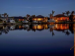Un port de plaisance au crépuscule, avec des bateaux amarrés, un bâtiment illuminé d’une lumière chaleureuse et des palmiers.
