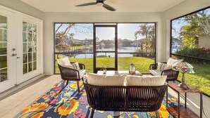 Una habitación con ventilador de techo, ventanas amplias, vistas a un lago y zona de estar exterior con un tapete colorido.