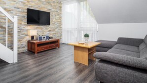 Un salon moderne avec un mur en pierre, un téléviseur à écran plat, une table basse en bois, un canapé gris et un escalier.