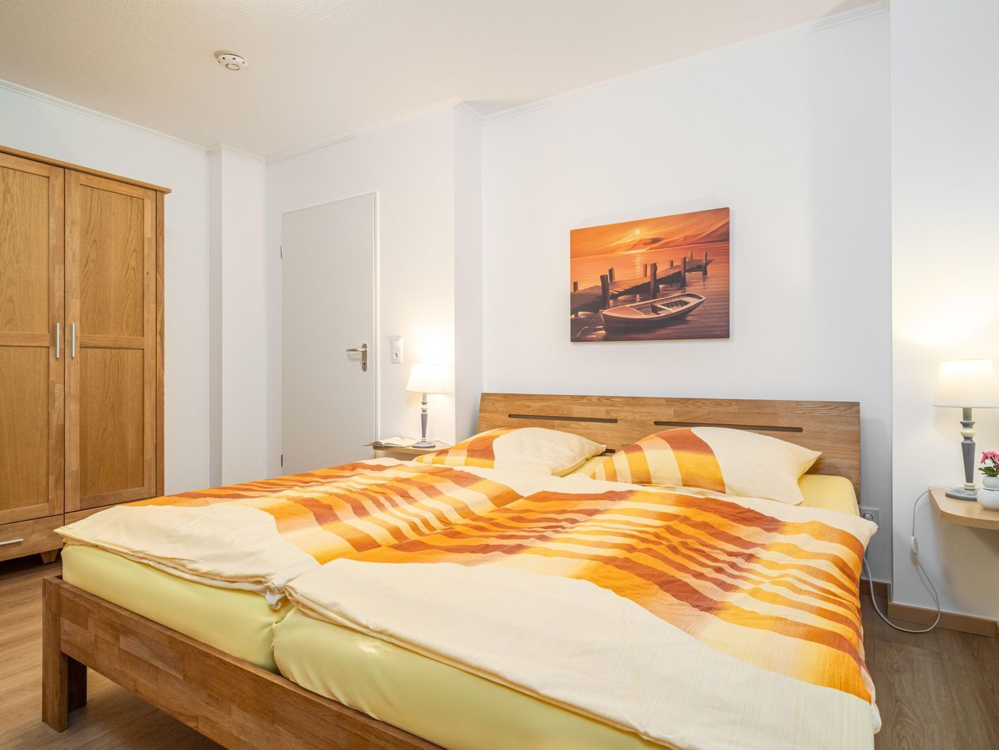 A bedroom with a wooden bed, a painting of a sunset over water, and a wooden wardrobe.