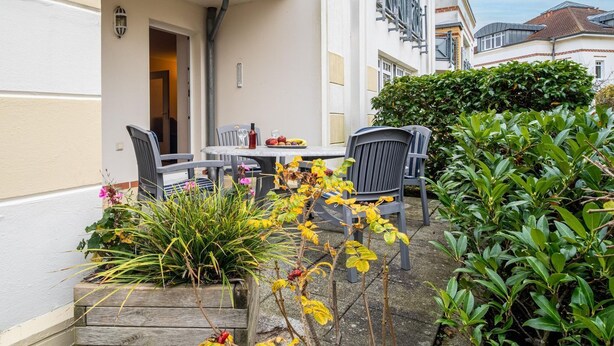 En udendørs terrasse med et bord og stole, omgivet af grøn vegetation og en bygnings facade.