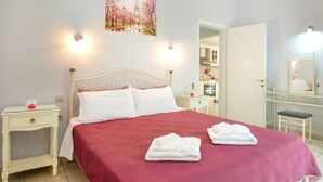 A neatly made bed with white linens, a red bedspread, and two folded towels on the bed. There is a white nightstand with a lamp and a small table with a mirror and a bench. A door leads to a bathroom with a mirror and a sink. - Relax near Corfu’s finest beaches in this charming villa for up to 4 guests (Halikounas)