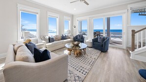 Ein geräumiges Wohnzimmer mit großem Fenster zum Meer, einem Deckenventilator und einer Holztreppe.