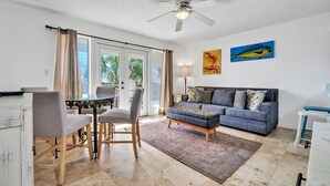 A living room with a sofa, a coffee table, a dining table with chairs, a ceiling fan, and a view through large glass doors.