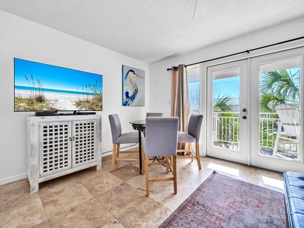 Una habitación con una mesa de comedor, sillas, un televisor en un mueble, un cuadro y vista a la playa a través de un ventanal corredera.