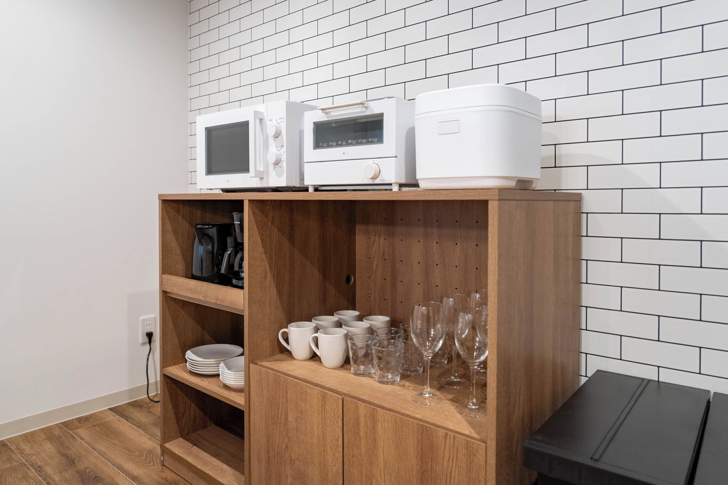 Een kitchenette met witte apparaten, een houten kastje met servies en glazen, en een zwarte tafel.