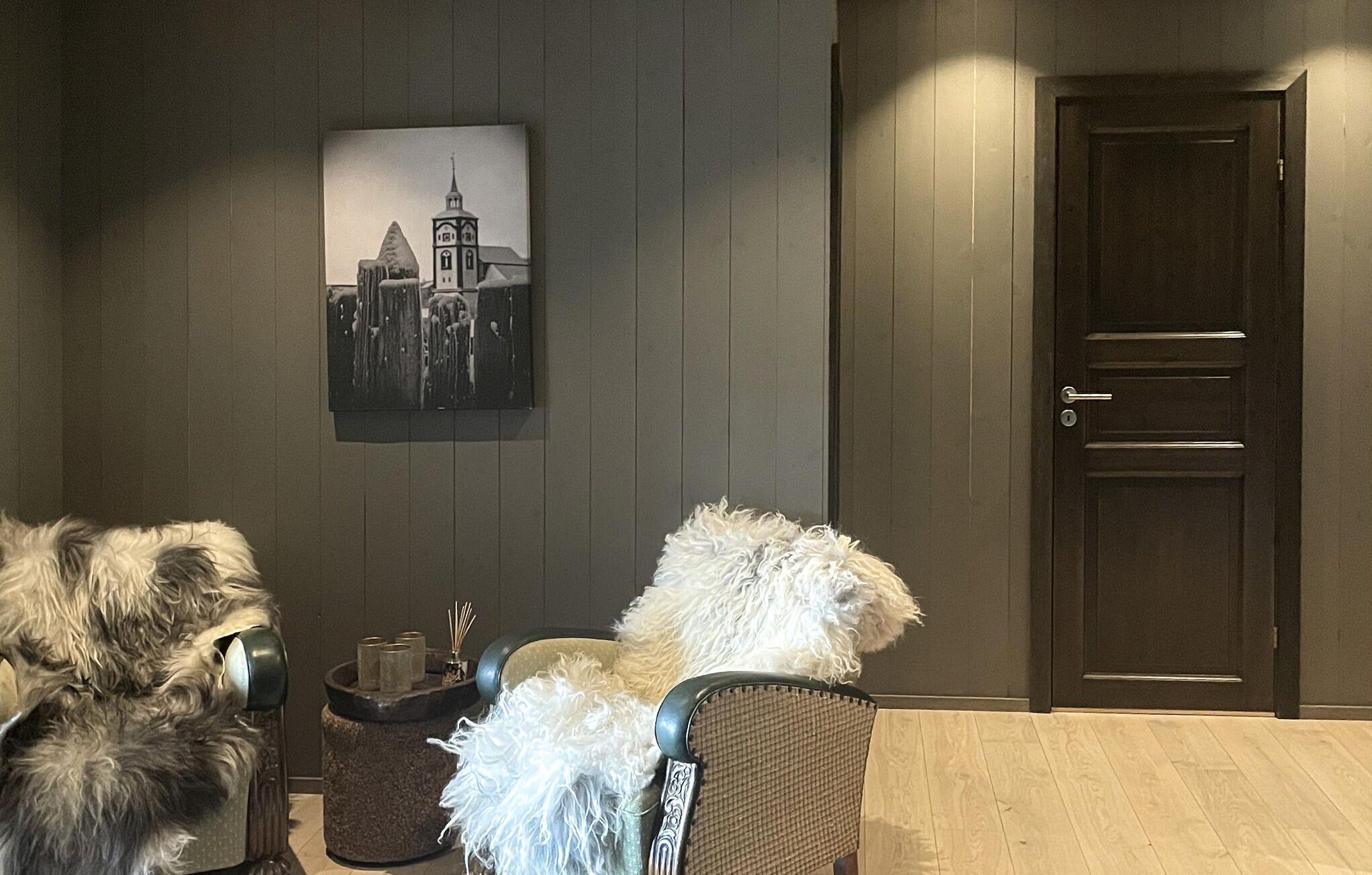 Ein Zimmer mit einer dunkelholzigen Tür, einem gerahmten Stadtbild an der Wand, einem flauschigen weißen Sessel und einem kleinen runden Tisch mit Kerzen.