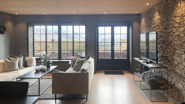 A modern living room with a stone wall, large windows, a sofa, a coffee table, and a TV.