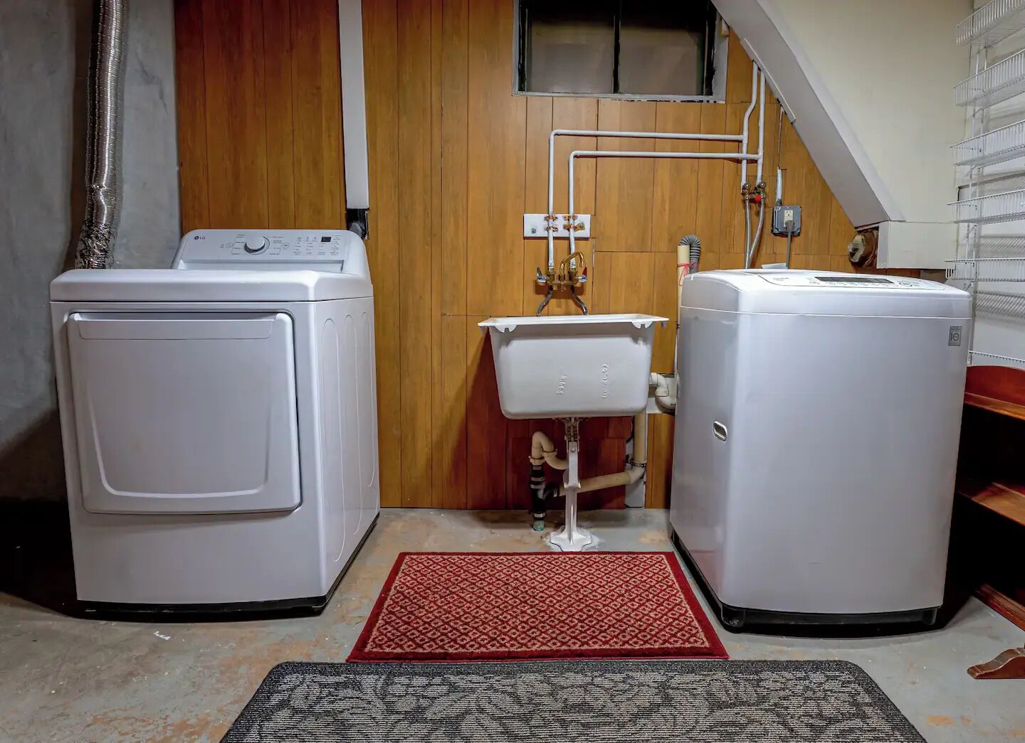 Een wasruimte met twee witte wasmachines, een rood-zwarte vloerkleed en een houten wand met een raam en sanitaire voorzieningen.