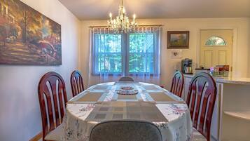 Une salle à manger avec une table dressée pour quatre personnes, entourée de chaises en bois, d’un lustre et d’une grande fenêtre ornée de rideaux.