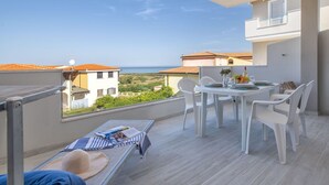 Un balcón con mesa y sillas, un banco con sombrero y revista, y vista al mar y edificios.