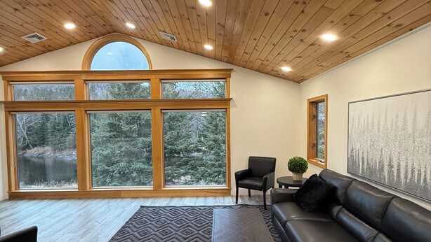 A spacious living room with a large window, a black leather sofa, a patterned rug, and a wooden ceiling. - Riverview Retreat! Near Trails! (Fort Kent)