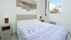 Un dormitorio con cama, mesita de noche, lámpara, ventana con cortinas y una obra de arte colgada en la pared.