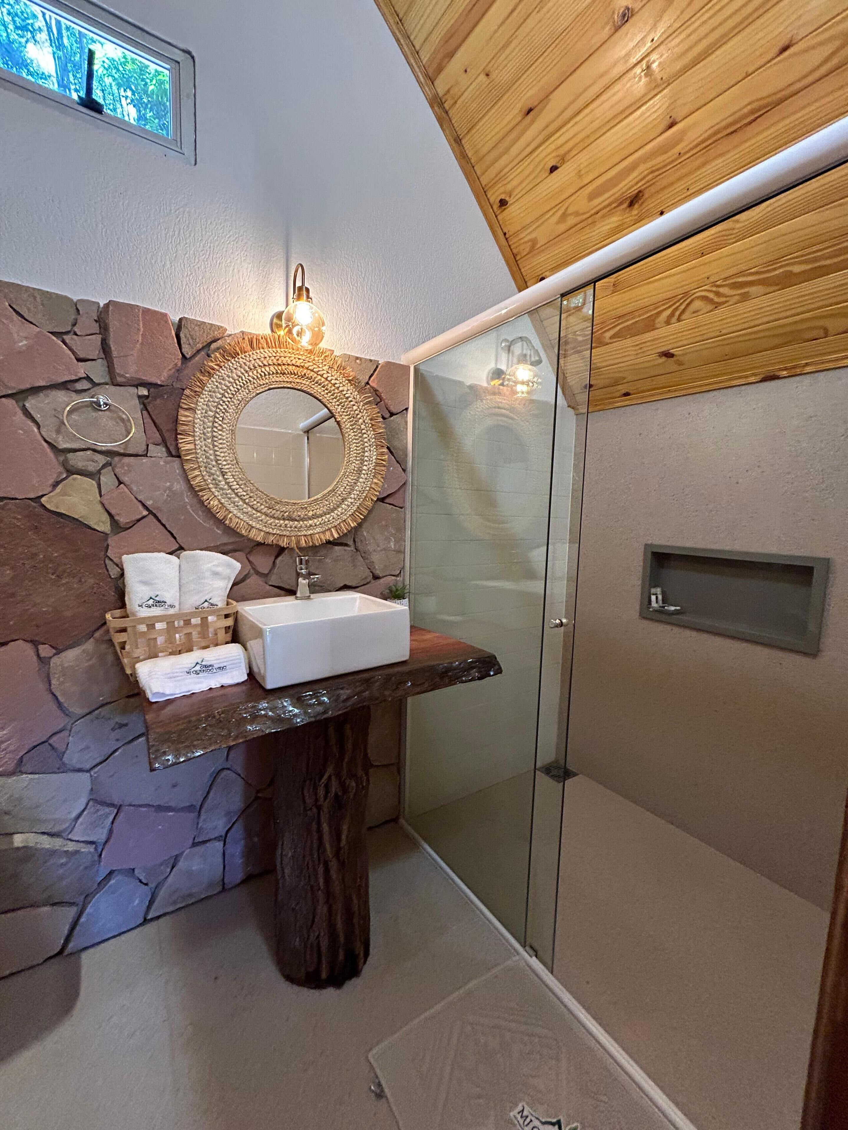 A bathroom with a stone wall, a wooden vanity, a round mirror, a glass shower enclosure, and a small shelf.