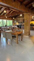 Un comedor amplio con mesas y sillas de madera, un pilar de ladrillo y una cocina al fondo.