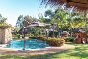 Zona de piscina con sombrillas, un pequeño quiosco y abundante vegetación.