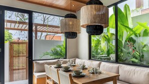 A dining area with a wooden table set for four, a sofa, and large windows with a view of greenery. - Nanna Villa Unit 2 by Betterplace (Canggu)