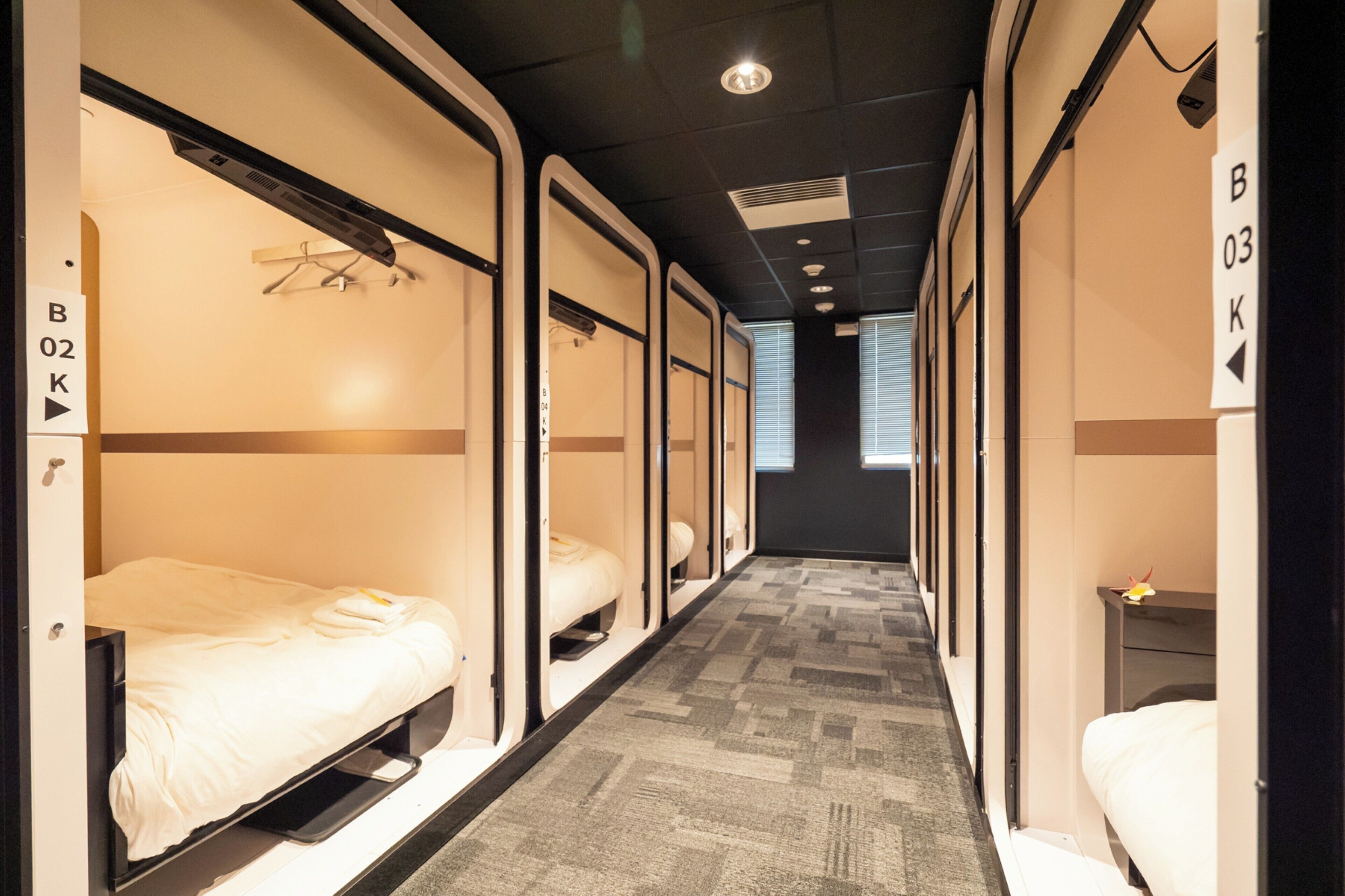 A corridor with bunk beds, each with a small table and a hanging rack. Signs indicate room numbers B02 and B03.
