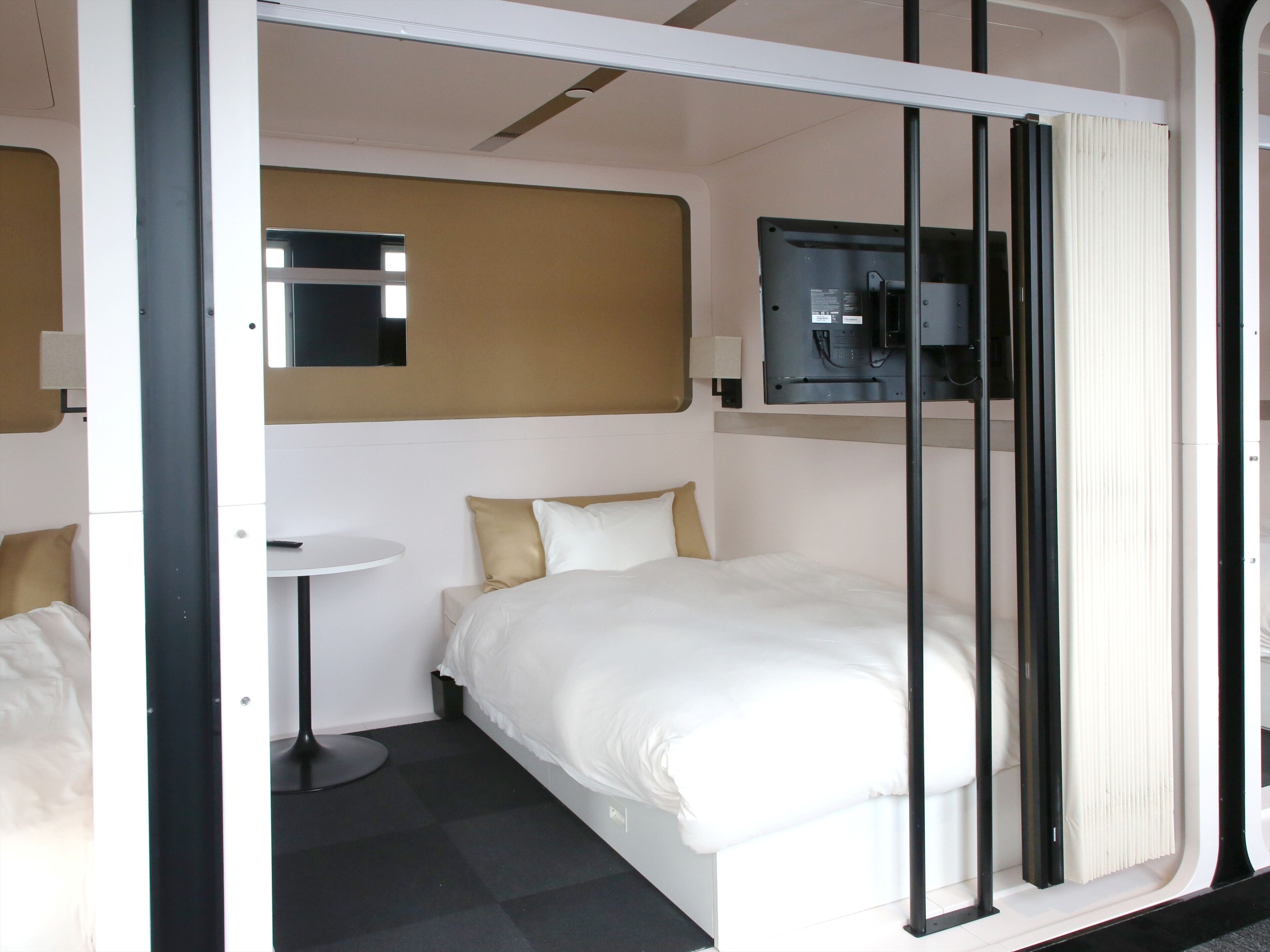 A modern hotel room with a bed, a small round table, a flat-screen TV mounted on the wall, and a window with curtains.