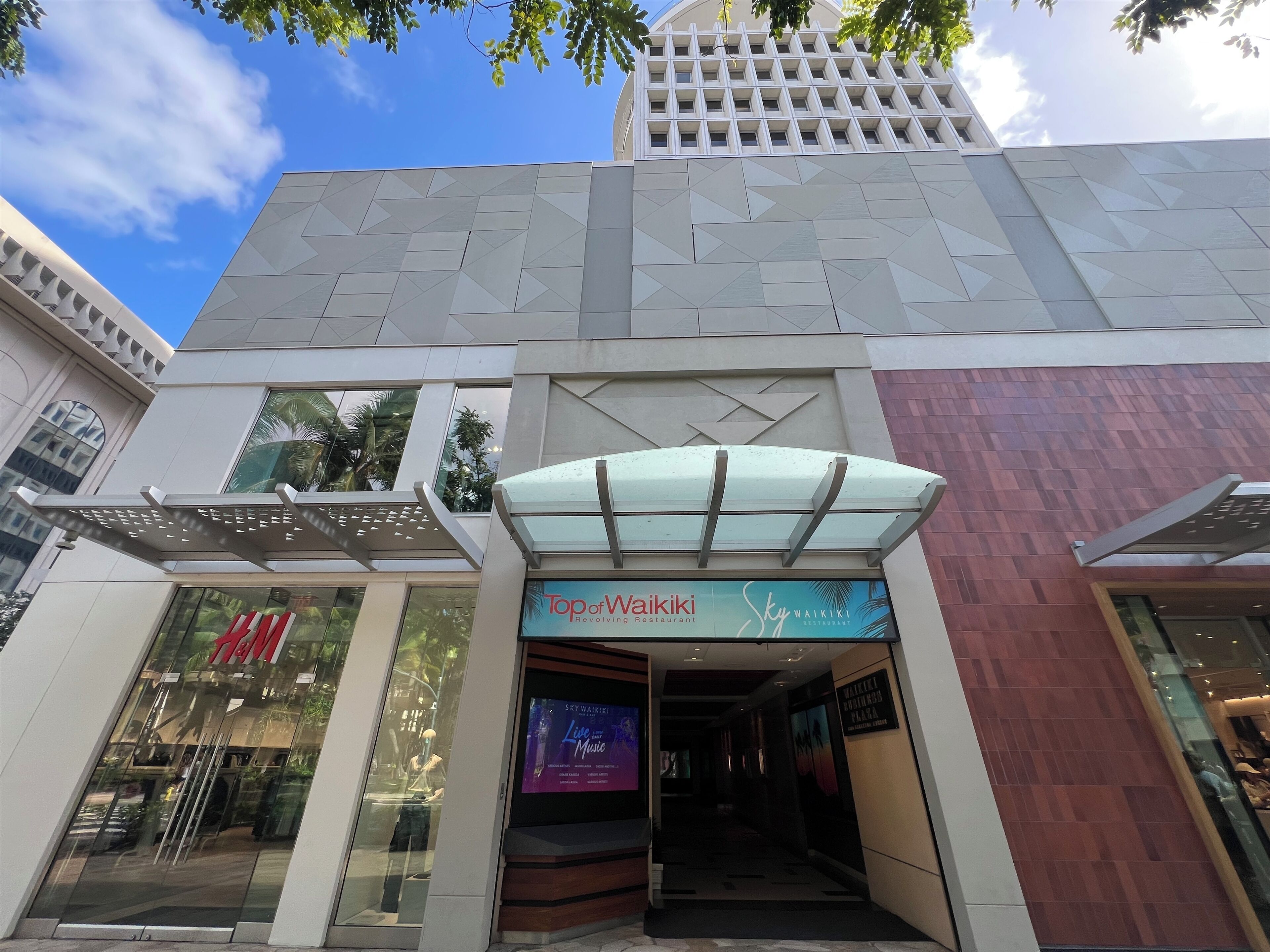 Un edificio moderno con entrada de vidrio, una tienda H&M a la izquierda y un letrero Sky Waikiki sobre la entrada.