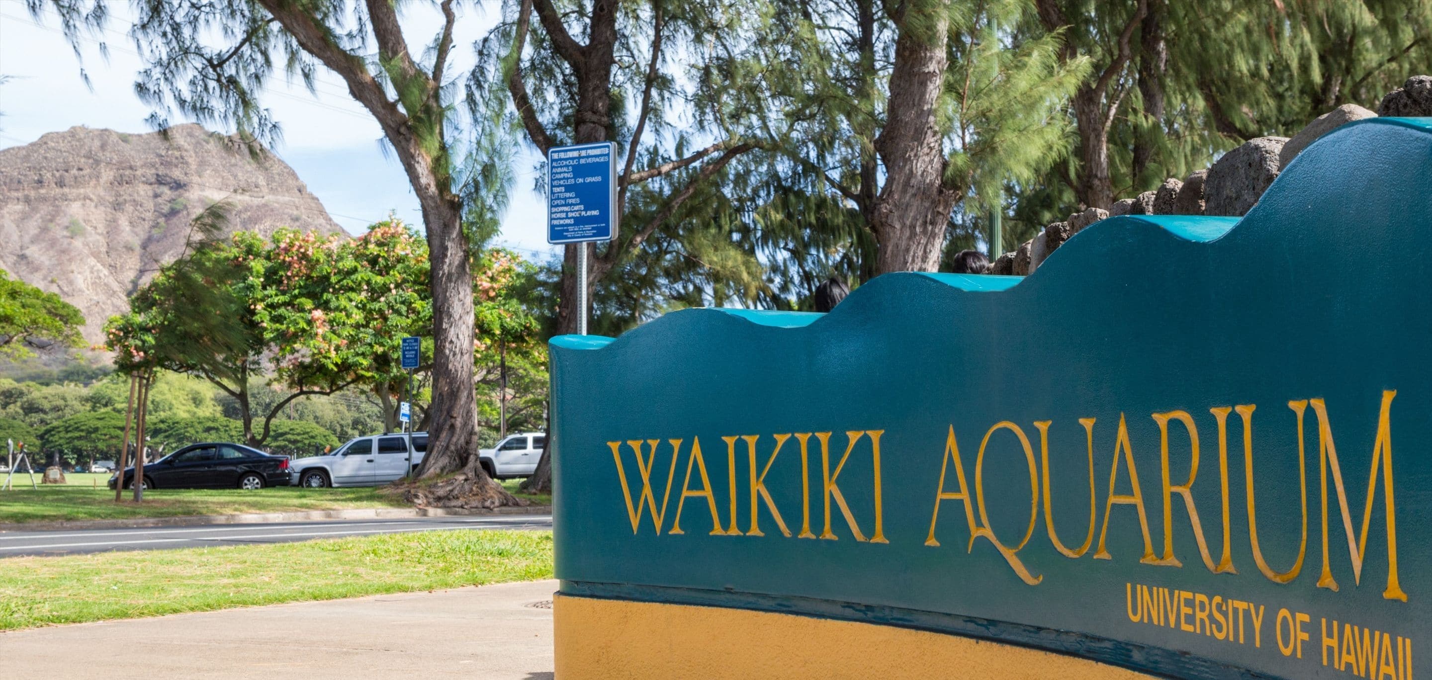 Señal del Acuario de Waikiki con montañas al fondo y coches estacionados.