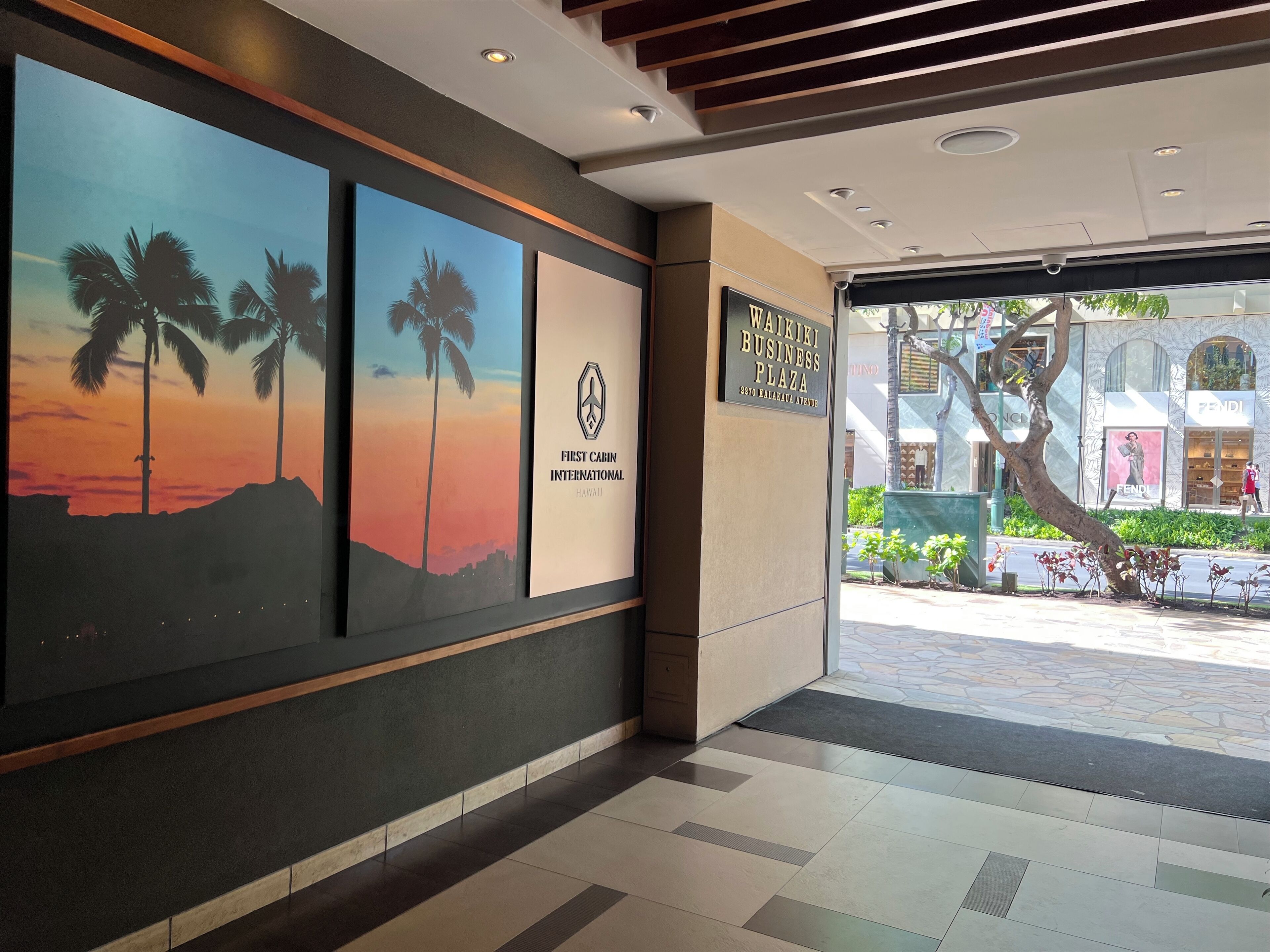 A hotel lobby with a reception desk, framed artwork depicting a sunset, and a sign for Waikiki Business Plaza.