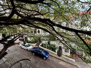 Straßenansicht mit zwei parkenden Autos, einem Gebäude mit rotem Dach und einem Baum mit Hängetöpfen.