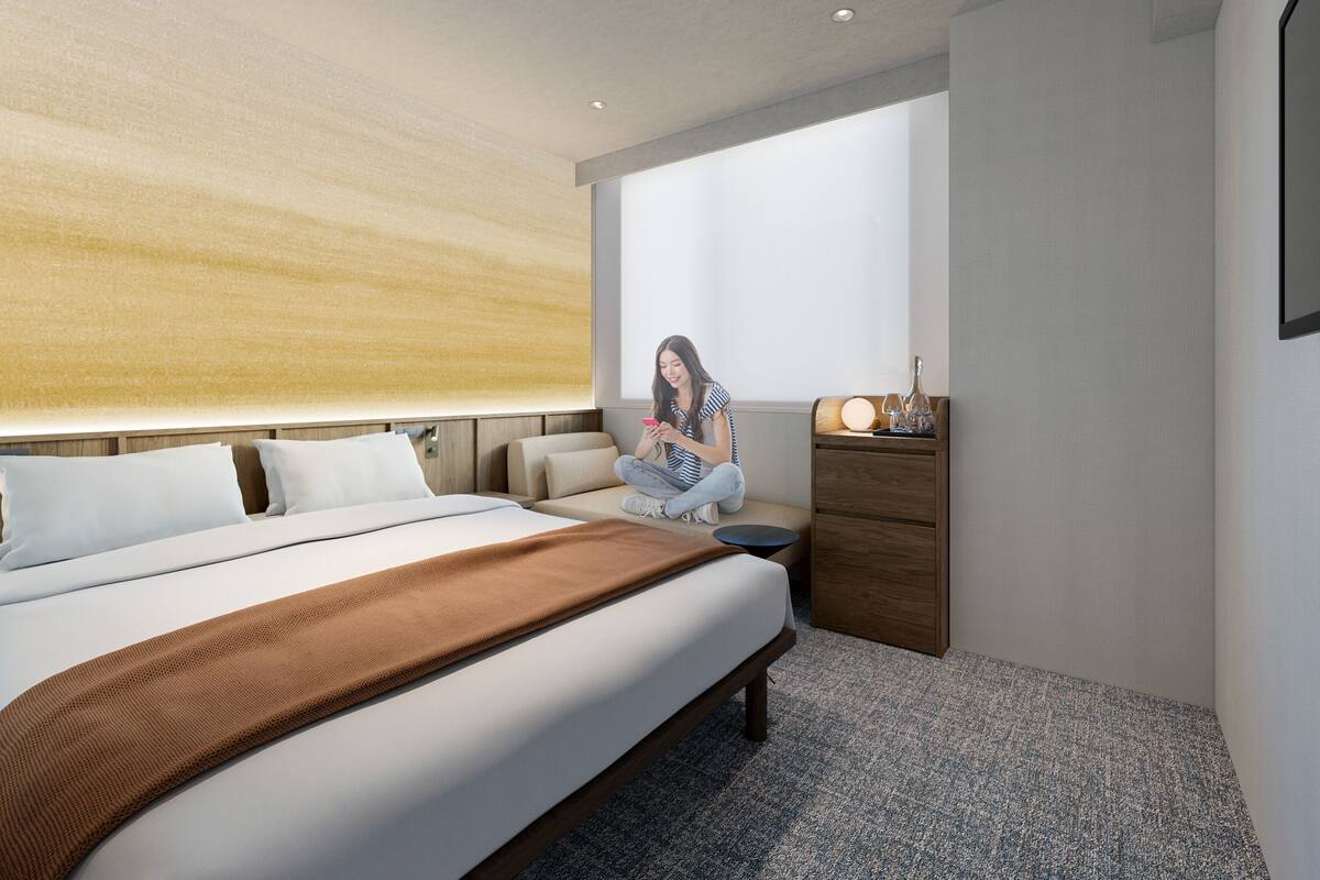 A modern hotel room with a large bed, a wooden headboard, a small seating area, and a nightstand with a lamp.